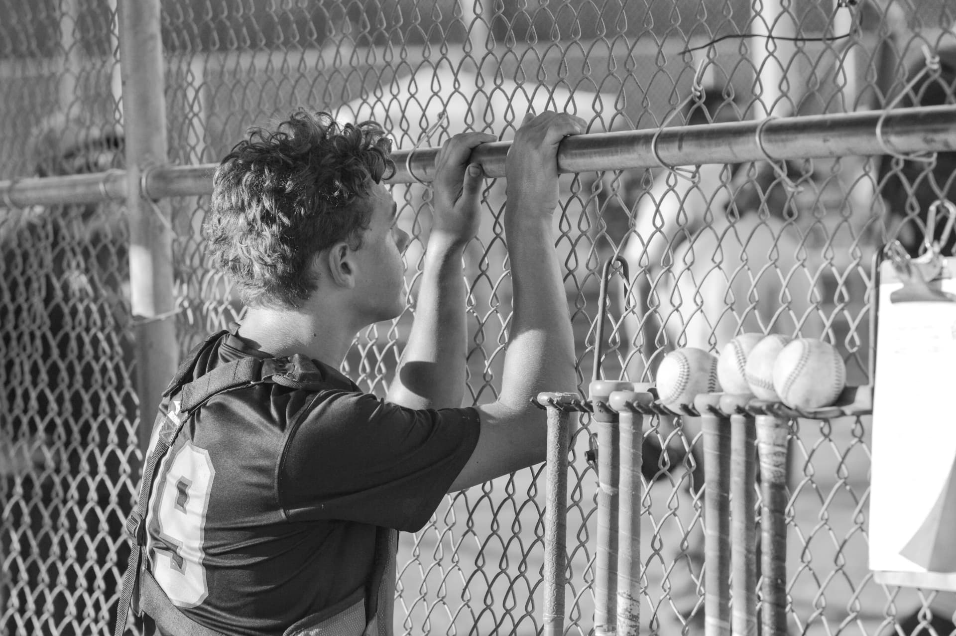 A young ballplayer leaning on the dugout fence beside bats and baseballs